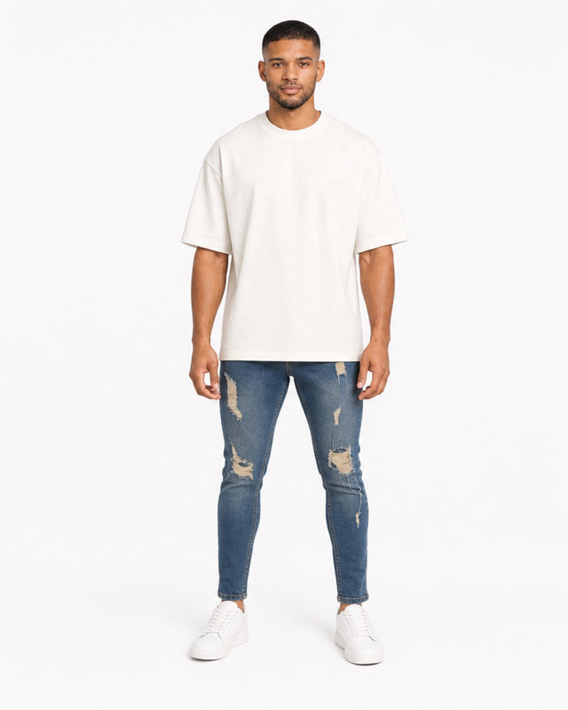 A man stands confidently in distressed blue jeans, paired with a loose-fitting white t-shirt and white sneakers.