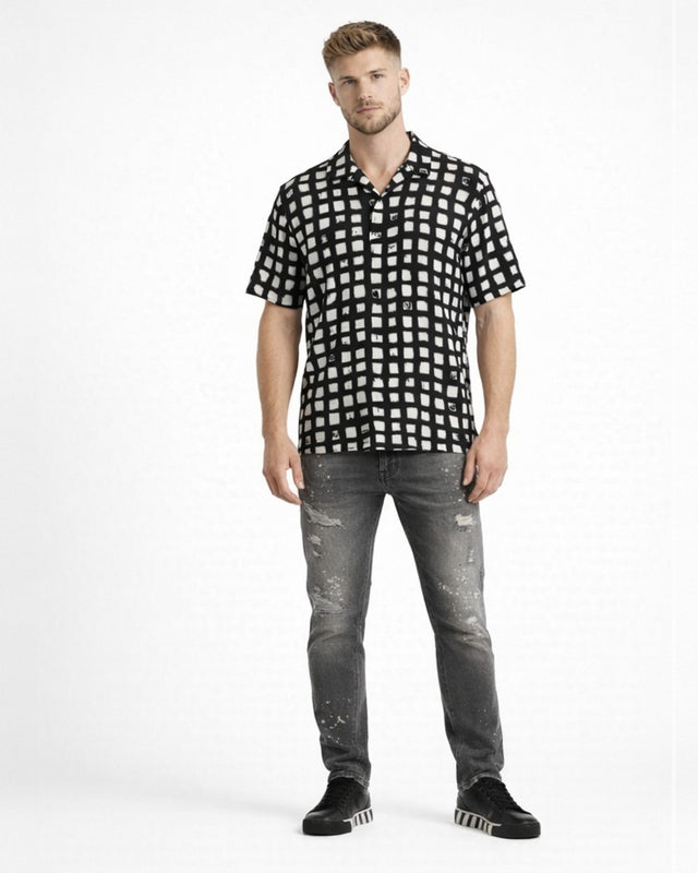 A man stands wearing a short-sleeve black and white checkered shirt paired with distressed gray jeans and black sneak...