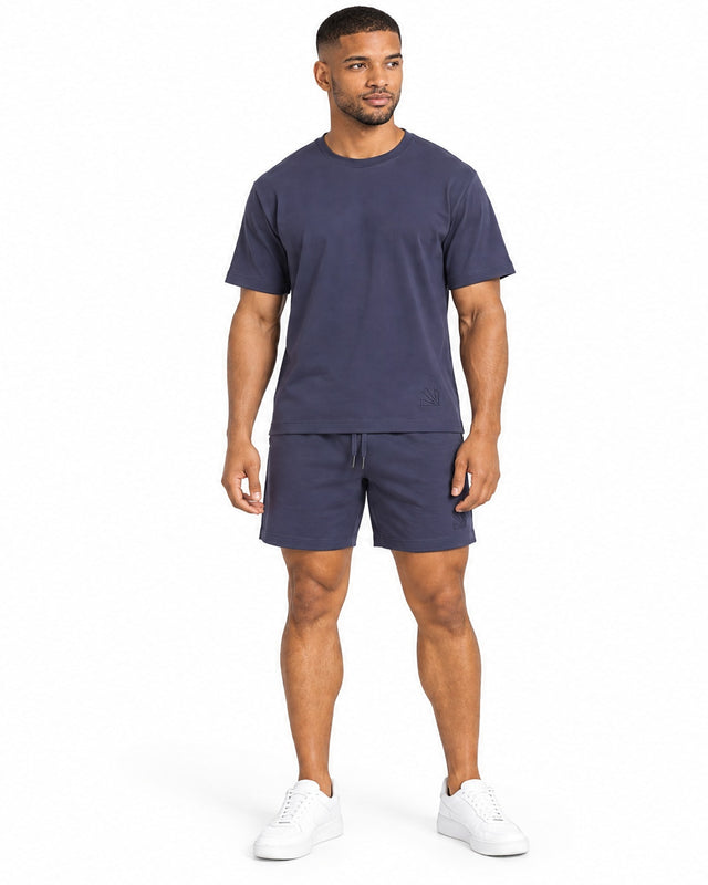 A man stands wearing navy shorts and a matching t-shirt, paired with white sneakers.
