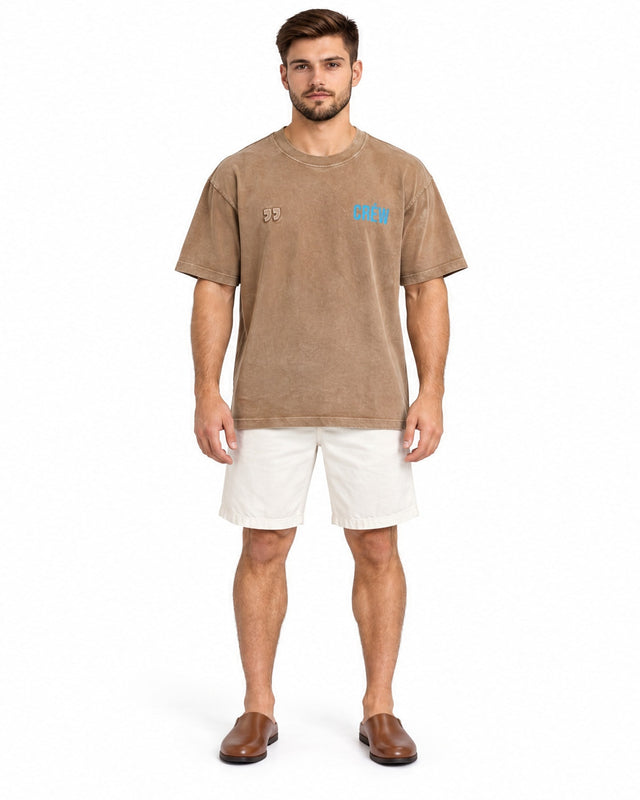 A man stands wearing a brown oversized t-shirt with blue text, paired with white shorts and brown slip-on shoes.