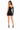 A woman stands in a fitted black dress with one long sleeve, showcasing a stylish back design and high heels.