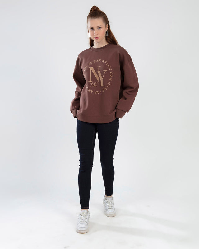Model wearing a brown NY graphic sweatshirt and black skinny jeans in a streetwear style studio shot.