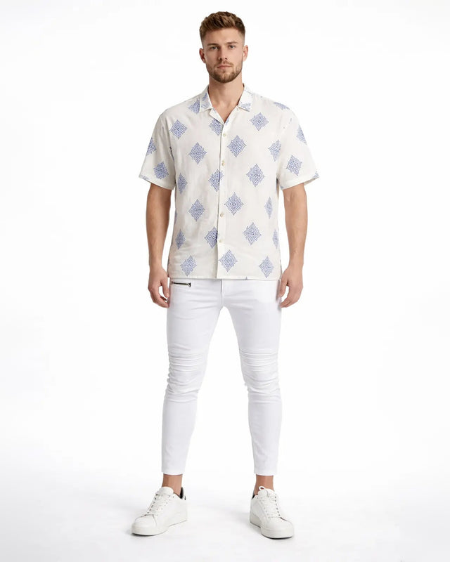 A man stands confidently in a short-sleeve patterned shirt, paired with white skinny jeans and sneakers.