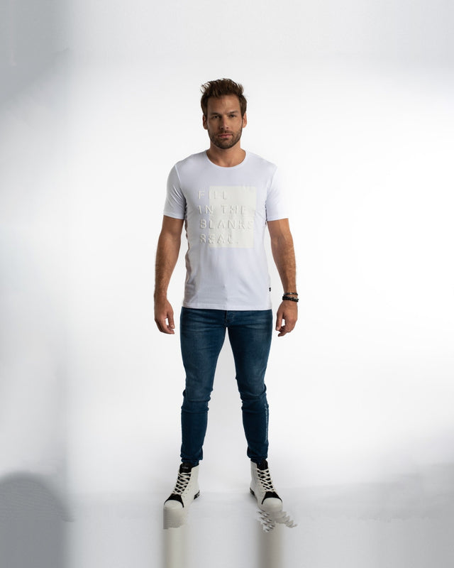 A man stands confidently wearing a white short-sleeve shirt with textured lettering, paired with blue jeans and styli...