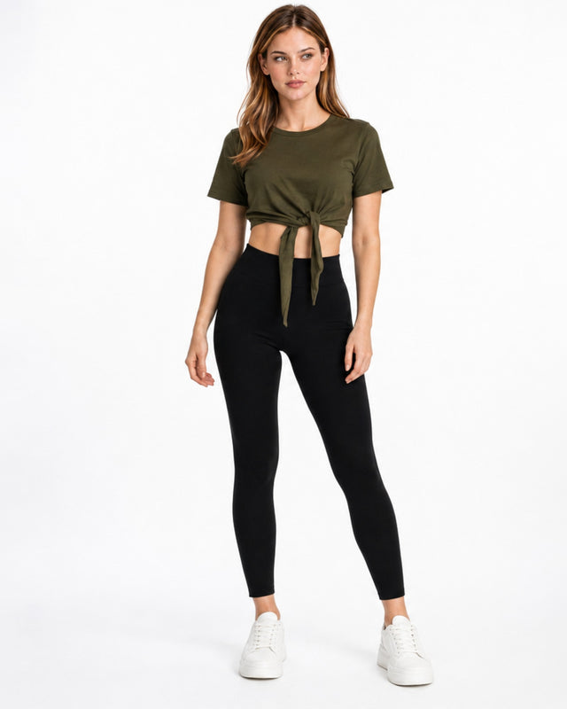A woman stands confidently in a cropped olive green top tied at the waist, paired with black leggings and white sneak...