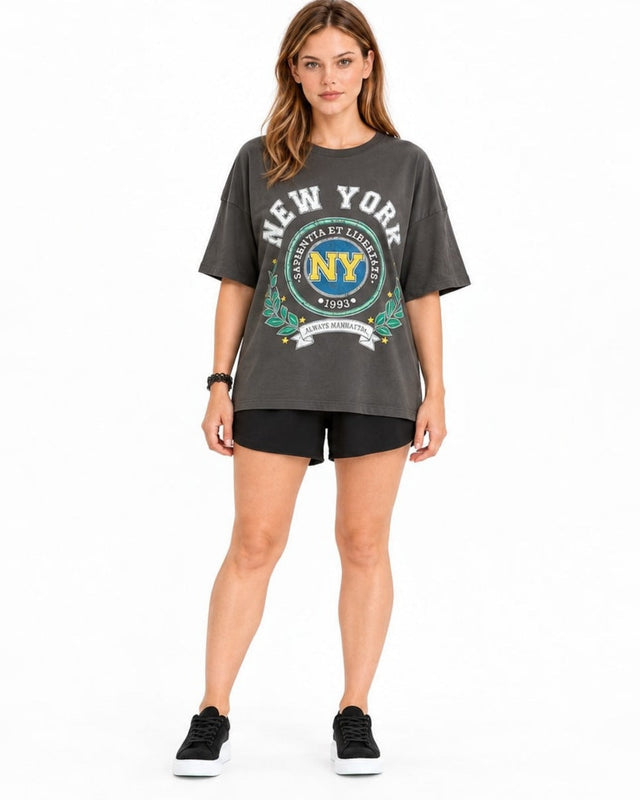 A woman stands wearing a loose-fitting gray t-shirt with a colorful New York graphic, paired with black shorts and sn...