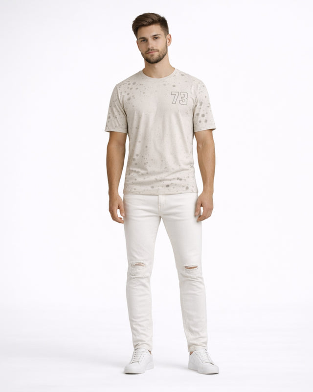 A man stands confidently in light-colored ripped jeans and a speckled t-shirt, paired with white sneakers.