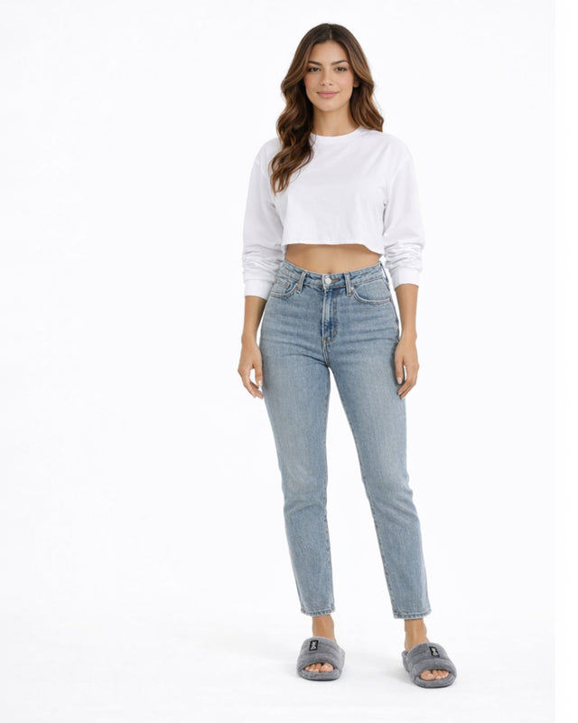 A woman stands confidently in a cropped white long-sleeve shirt and light blue jeans, paired with gray slippers.