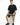 A man sits on a white pedestal wearing a black oversized t-shirt and dark shorts, paired with black sneakers.