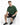 A man sits on a white cube wearing a green oversized t-shirt, black shorts, and black sneakers with white stripes.