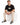 A man sits on a white cube wearing beige jogger pants, a black t-shirt, and stylish black sneakers.