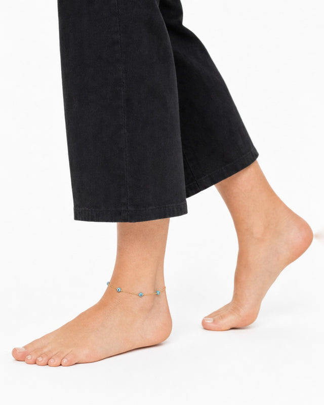 A close-up of a bare foot with a delicate gold anklet featuring blue decorative beads.