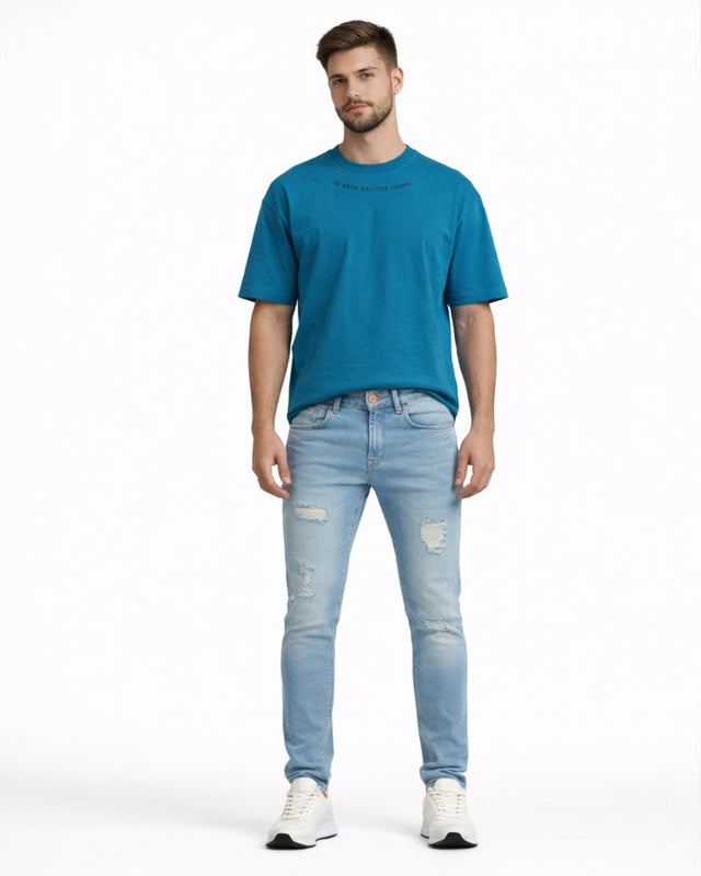 A man stands wearing light blue distressed jeans and a teal t-shirt, paired with white sneakers.