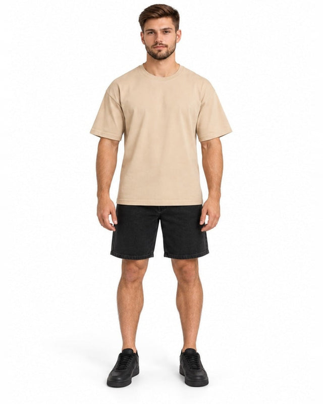 A man stands confidently wearing a beige short-sleeve t-shirt paired with black denim shorts and sneakers.