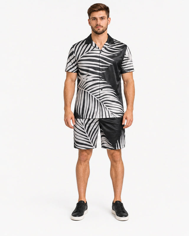 A man stands wearing a short-sleeve shirt and matching shorts with a black and white palm leaf pattern.