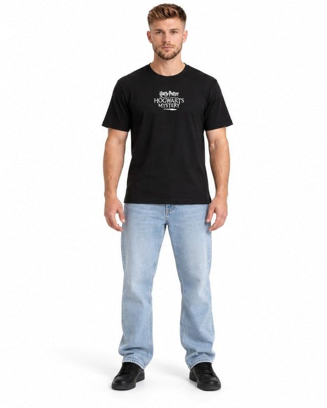 A man stands wearing a black t-shirt with a graphic design, paired with light blue jeans and black shoes.