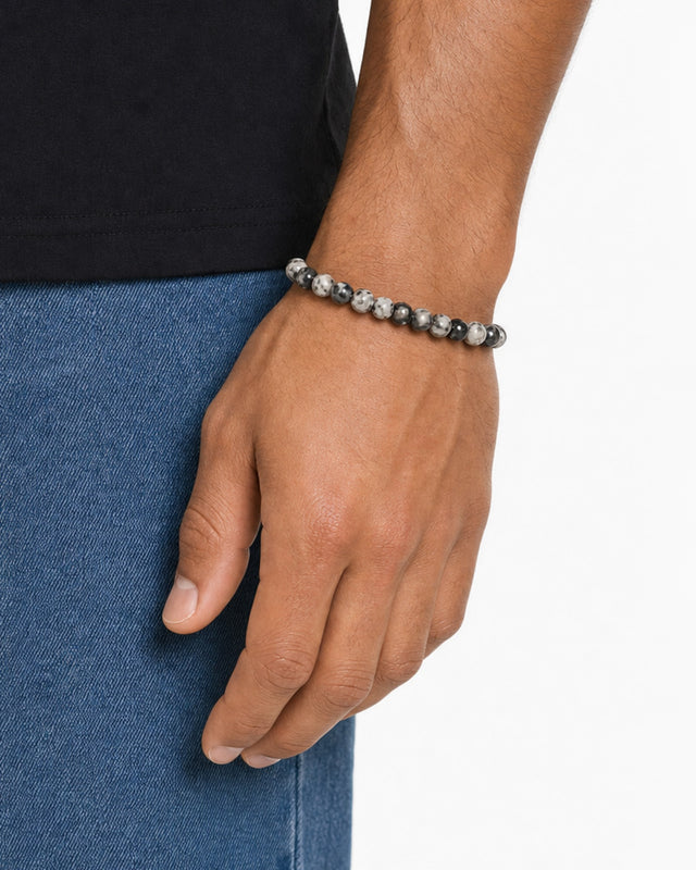 A close-up of a hand wearing a beaded bracelet with a mix of gray and black stones, resting on denim.