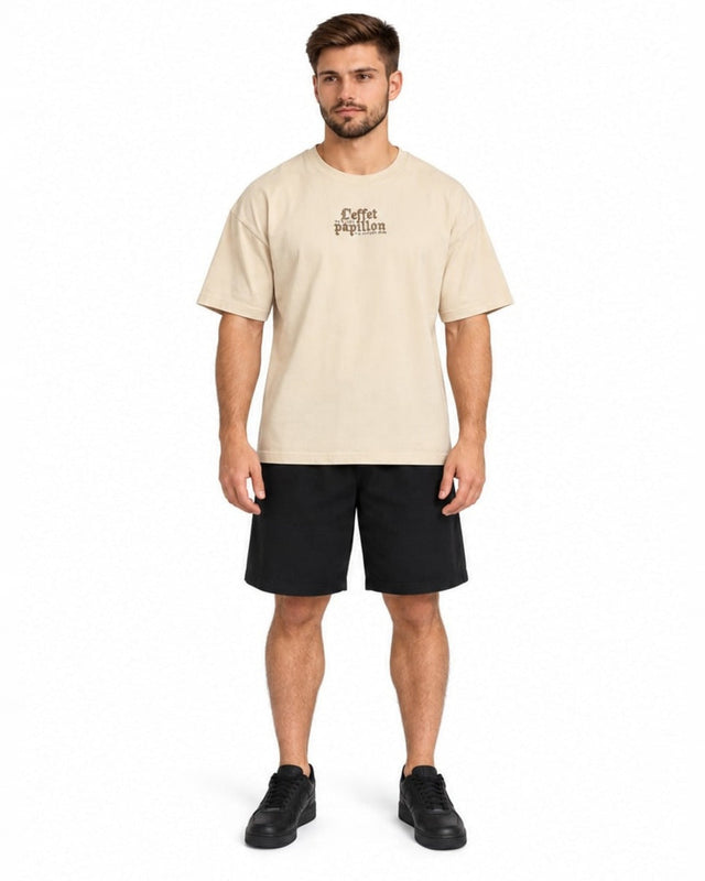 A man stands wearing a beige oversized t-shirt with a graphic design, paired with black shorts and sneakers.