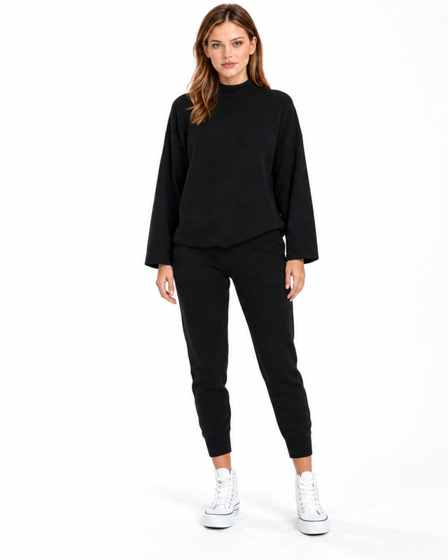 A woman stands in a relaxed pose wearing a black top and matching sweatpants, paired with white sneakers.