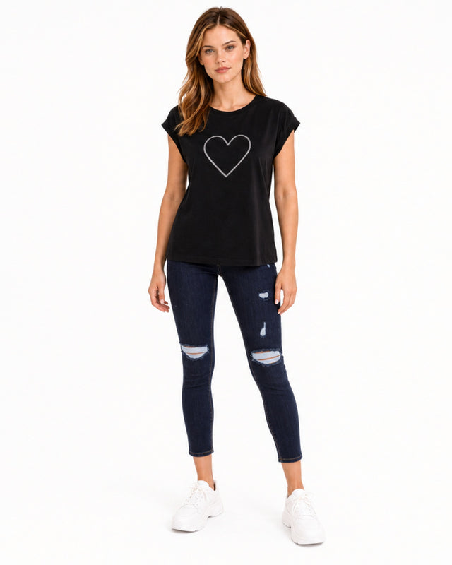 A woman stands in a black short-sleeve top with a heart graphic, paired with distressed dark jeans and white sneakers.