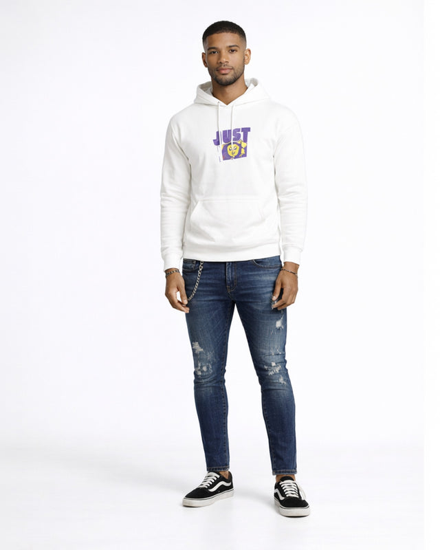 A man stands confidently in a white hoodie with a graphic design, paired with distressed blue jeans and black sneakers.
