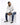 A man sits on a white pedestal wearing a light hoodie, distressed jeans, and black sneakers, looking thoughtfully to...