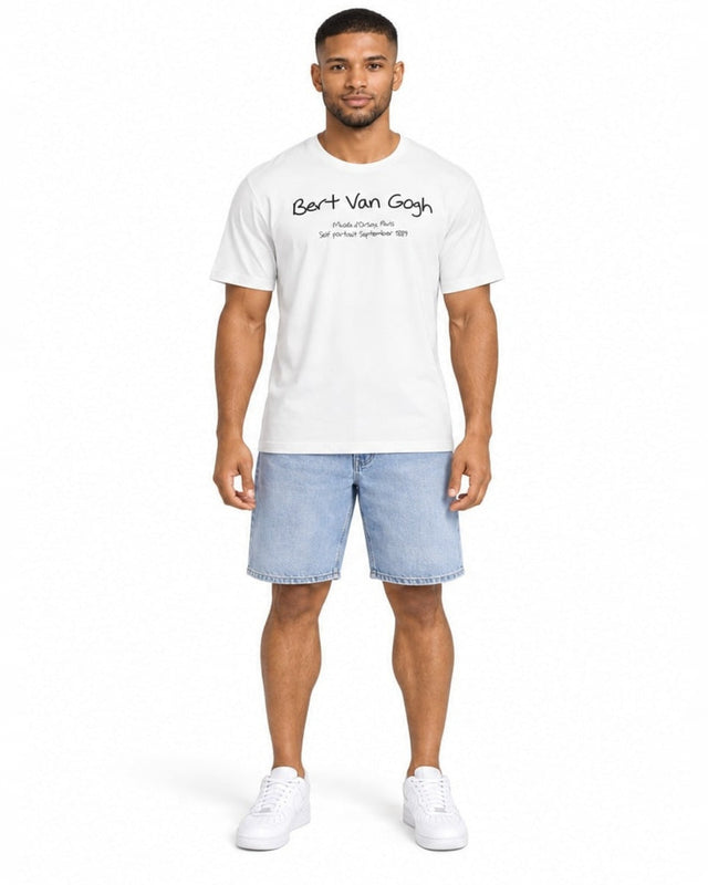 A man stands wearing a white t-shirt with artistic text and denim shorts, paired with white sneakers.