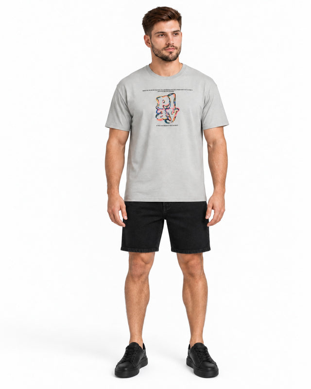 A man stands wearing a light gray t-shirt with a colorful graphic design, paired with black shorts and sneakers.