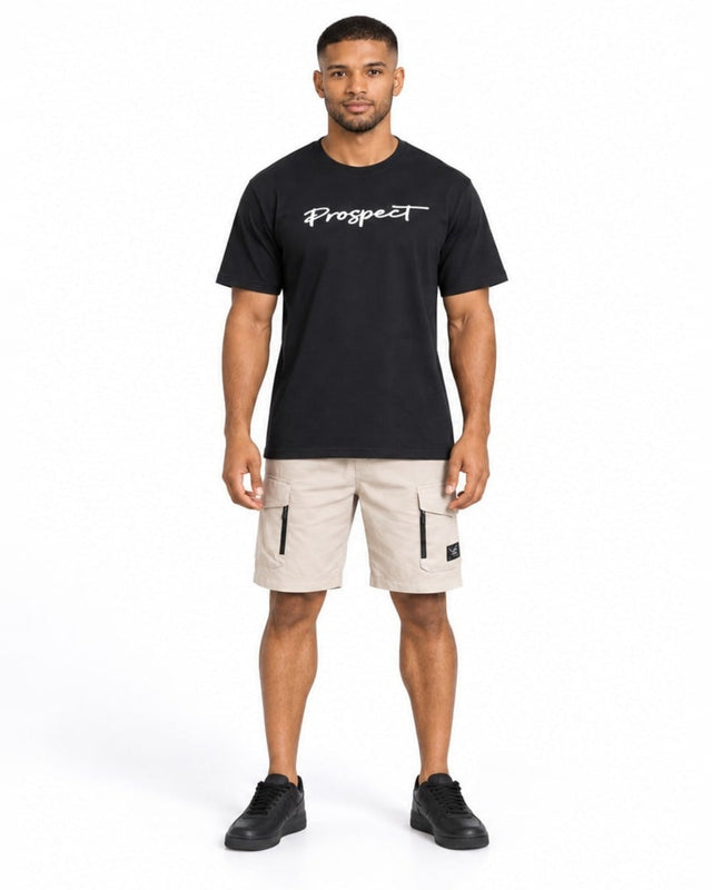 A man stands wearing a black t-shirt with white text, paired with beige cargo shorts and black sneakers.