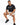 A man sits on a white stool wearing a black t-shirt and beige shorts, paired with black sneakers.