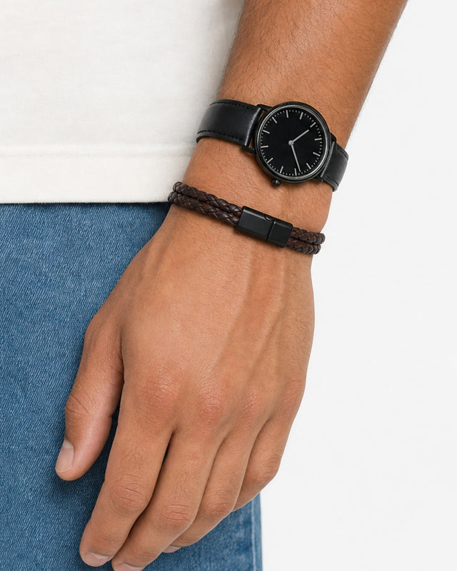 A close-up of a hand wearing a braided brown bracelet alongside a black watch on a denim background.