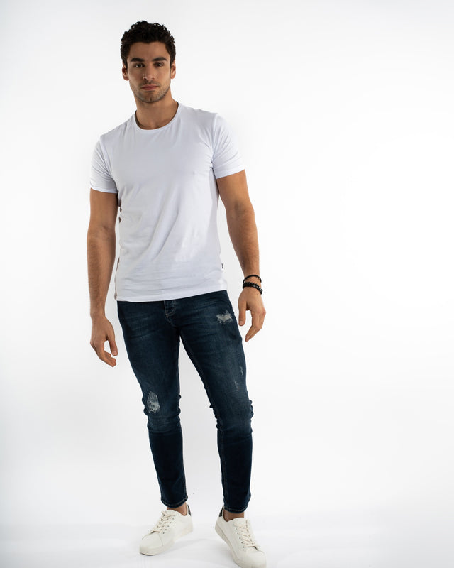 A man stands confidently in a fitted white t-shirt and distressed dark jeans, paired with white sneakers.