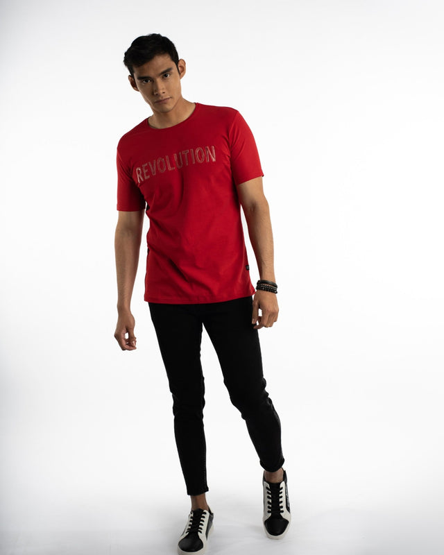 A young man poses in a red short-sleeve shirt with the word "REVOLUTION" printed on the front, paired with black jean...