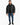 A man stands wearing a black puffer coat, black shirt, distressed jeans, and sneakers against a white background.