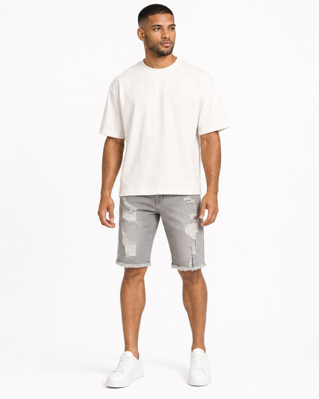 A man stands wearing a loose-fitting white t-shirt and distressed gray denim shorts, paired with white sneakers.