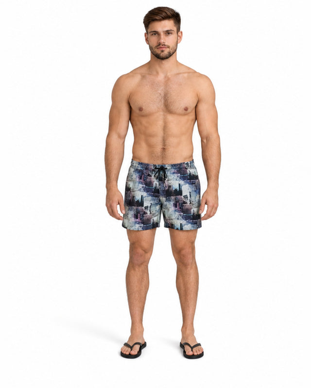 A shirtless man stands in colorful swim trunks with a cityscape pattern, wearing flip-flops.