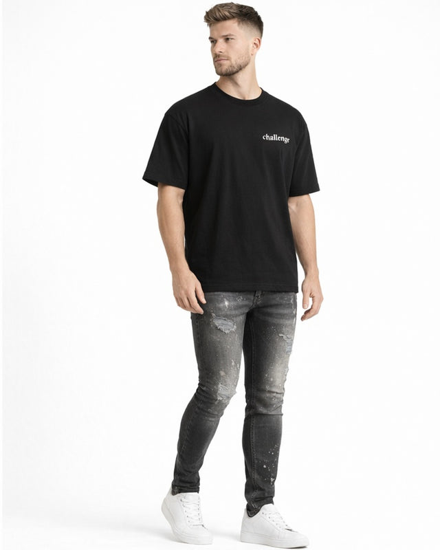 A man stands wearing a black t-shirt and distressed gray jeans, paired with white sneakers.