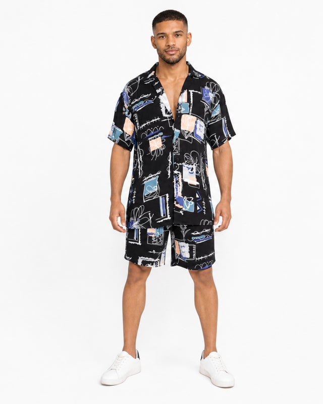 A man stands wearing a short-sleeve, patterned button shirt and matching shorts, paired with white sneakers.