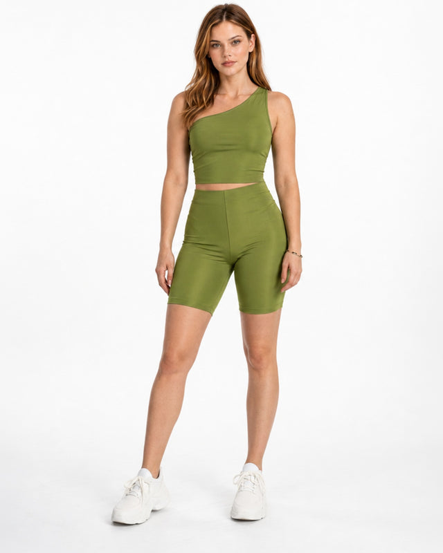 A model poses in olive green fitted shorts and a matching one-shoulder crop top, paired with white sneakers.