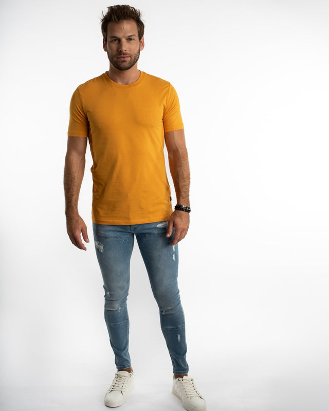 A man stands confidently in a fitted orange shirt paired with distressed blue jeans and white sneakers.