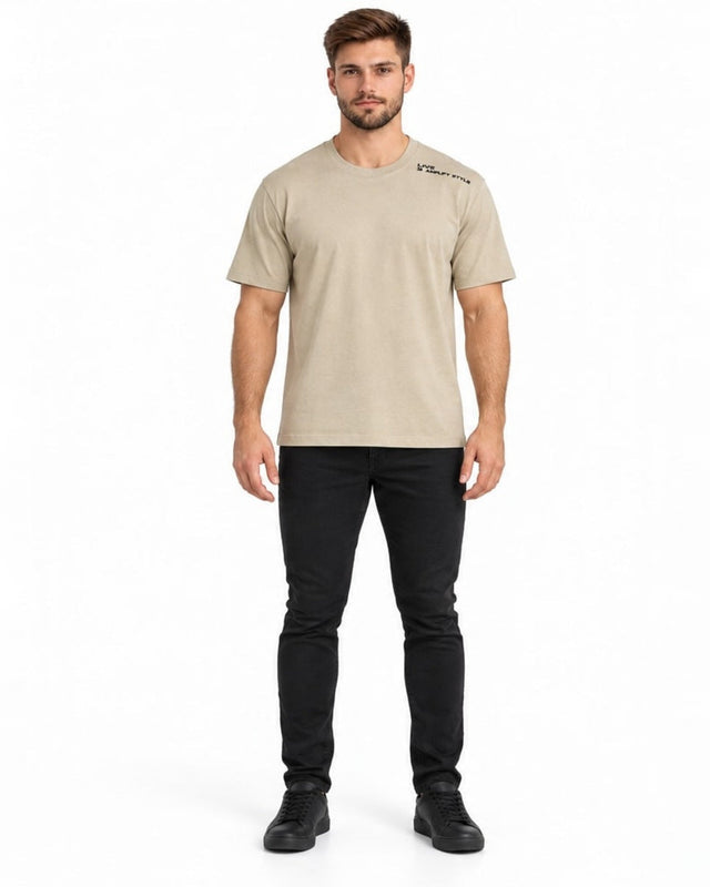 A man stands in a neutral-toned t-shirt paired with black jeans and sneakers, showcasing a casual style.