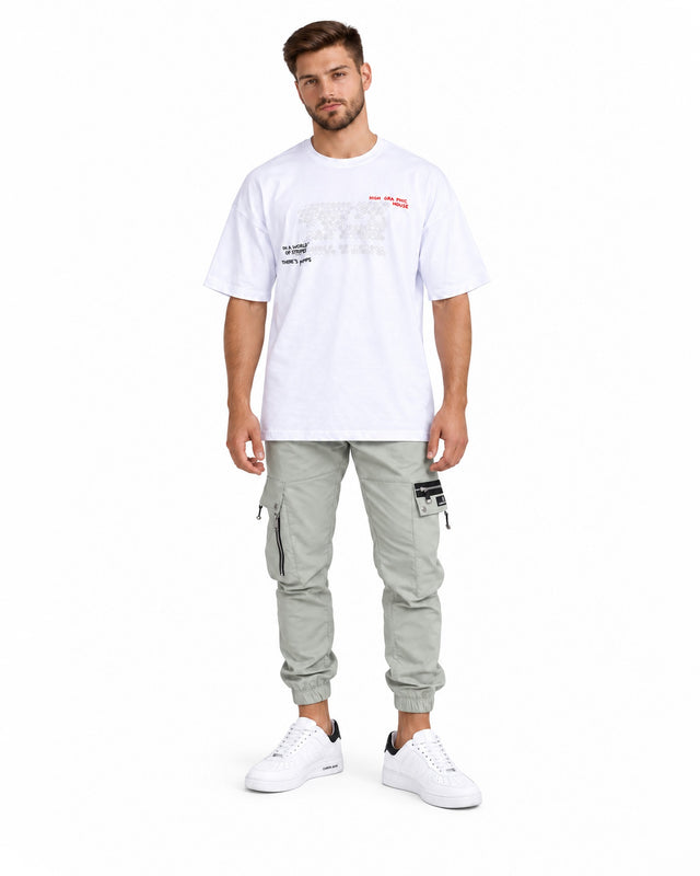 A man stands wearing a white graphic t-shirt, light green cargo pants, and white sneakers with black accents.