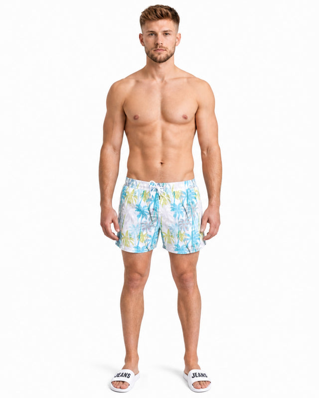A shirtless man stands in colorful swim trunks featuring a tropical palm tree print, wearing slide sandals.