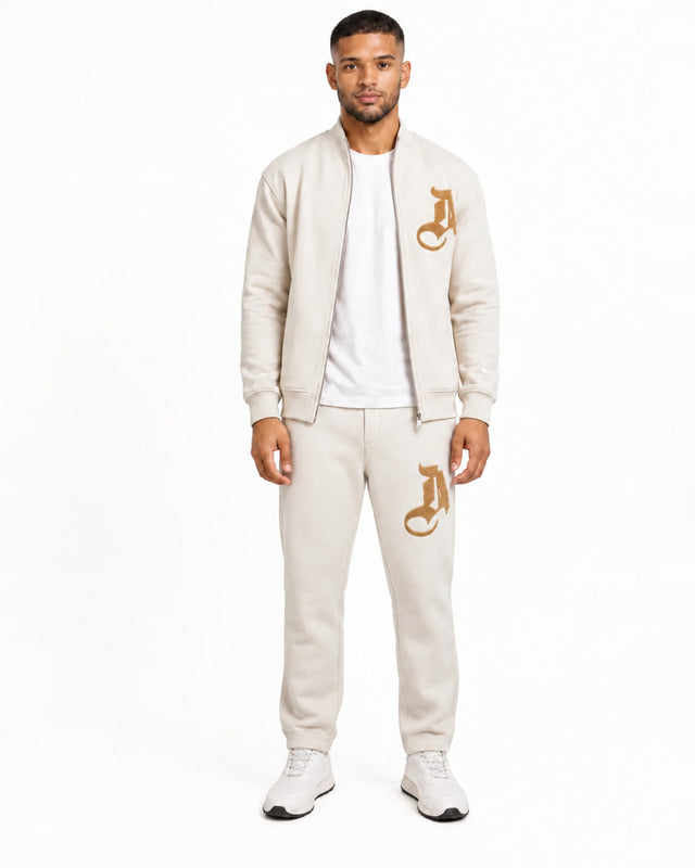 A man stands confidently in a light beige zip-up jacket and matching sweatpants, featuring a gold logo.