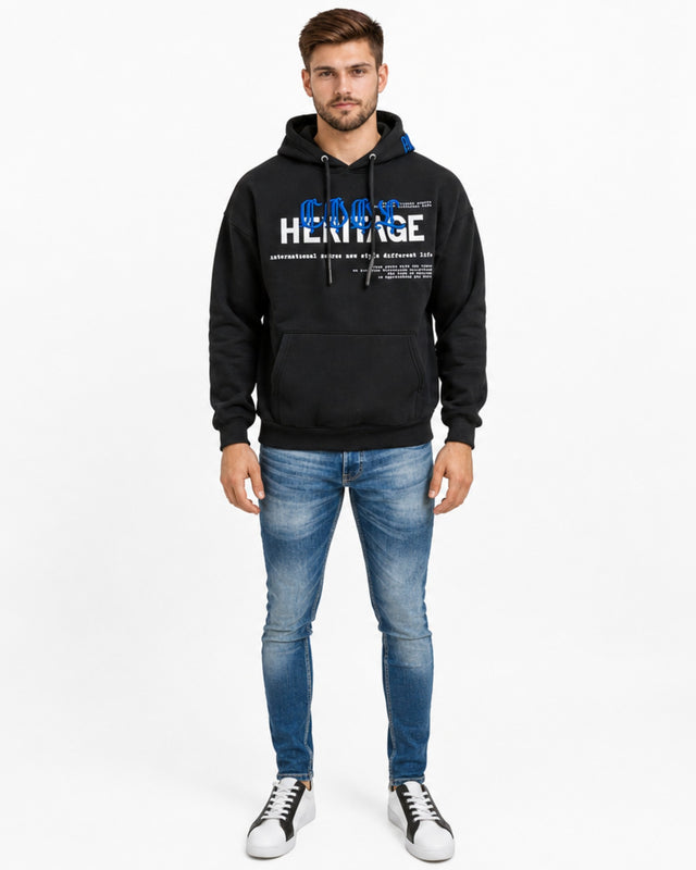 A man stands wearing a black hoodie with graphic text, paired with light blue jeans and stylish sneakers.