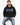 A man sits on a stool wearing a black hoodie with graphic text and blue accents, paired with blue jeans.