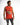 A man wearing a sleeveless bright orange top with graphic prints and distressed gray jeans, posing against a white ba...