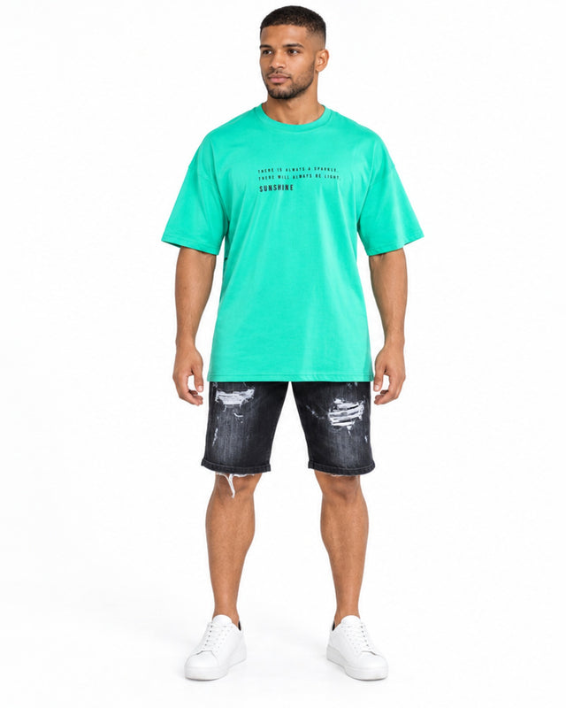 A man stands wearing a bright green oversized t-shirt with a graphic design, paired with distressed black shorts and...