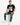 A man sits on a white cube, wearing a black t-shirt with a metallic graphic and distressed blue jeans.