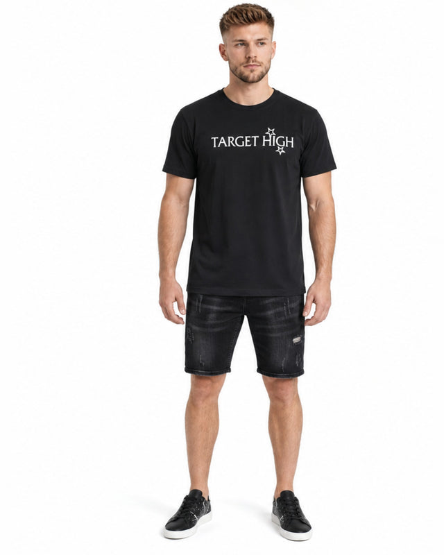 A man stands wearing a black t-shirt with a graphic design, paired with distressed denim shorts and sneakers.